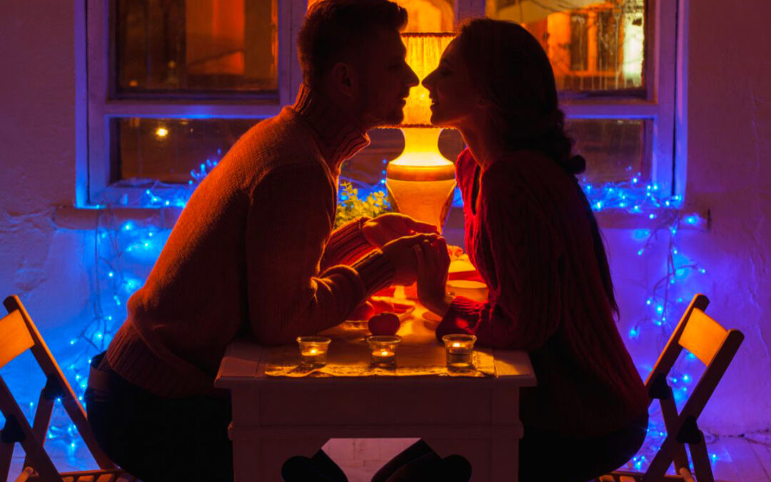 Un couple d'amoureux assis à une petite table dans la pénombre, éclairée par des lumières bleues et orangées. Une guirlande électrique bleue ajoute une ambiance douce et romantique, tandis que des bougies sur la table apportent une lueur tamisée. Ils se tiennent les mains et se penchent l'un vers l'autre, prêts à s'embrasser, capturant un moment d'intimité et de connexion passionnée.