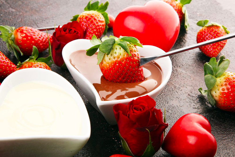 Une fondue au chocolat avec une fraise plantée sur une pique, trempée dans un bol de chocolat noir en forme de cœur. Autour, des fraises dispersées, quelques roses, et un autre ravier en forme de cœur rempli de chocolat blanc, créant une scène gourmande et romantique.