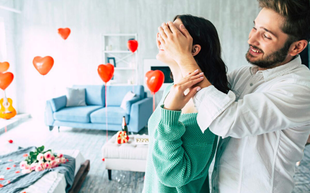 Un couple amoureux, l'homme cachant les yeux de sa femme avec ses mains, créant une scène ludique et romantique. En arrière-plan, un salon décoré pour la Saint-Valentin avec des ballons en forme de cœur suspendus, un bouquet de roses roses posé sur le coin du canapé, ainsi qu'une bouteille et une boîte de chocolats en forme de cœur et d'autres friandises disposées sur un autre coin, ajoutant une touche de douceur et de festivité à l'ambiance.